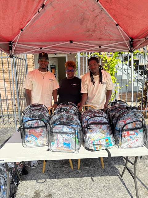 Three people standing behind a table under a red tent canopy displaying four colorful backpacks filled with supplies