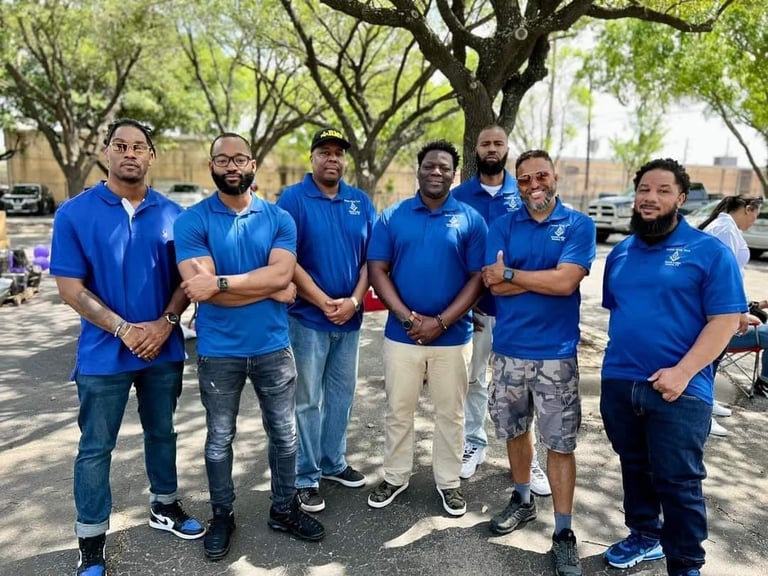 Group of eight men wearing matching blue shirts standing together outdoors under trees in a parking lot