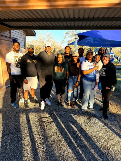 Group of diverse people standing together under a blue umbrella at an outdoor event with bare trees in background