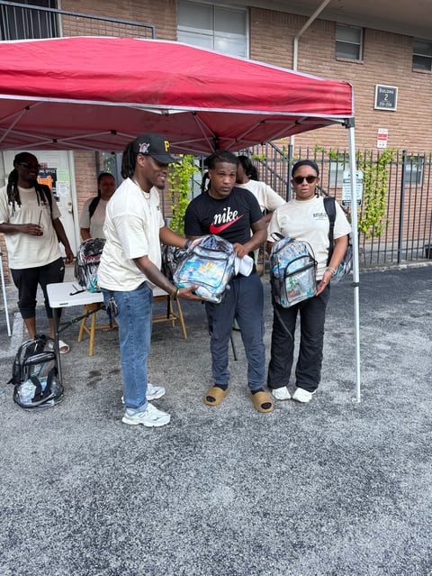Group of people distributing backpacks under a red canopy at an outdoor community event