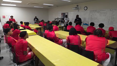 Group of people in pink and red shirts seated at long yellow tables in a classroom during a training or meeting session