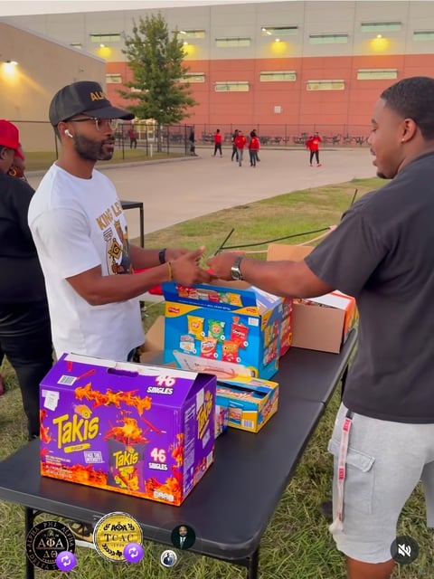 Two men exchange colorful snack boxes at a table outside a school building with people playing in the background