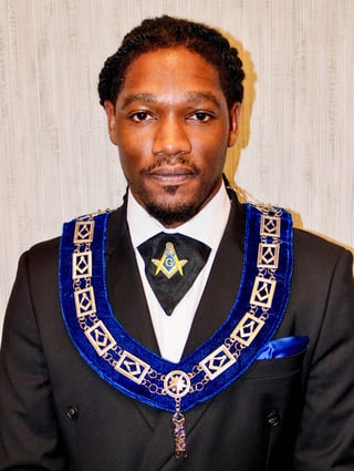 Man in formal black suit wearing blue and gold Masonic ceremonial regalia with emblems and chain