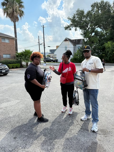 Three people stand in a parking lot, one holding a bicycle. The woman in the red shirt appears to be receiving or discussing something with the others in a community outdoor setting.