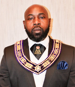 Man in formal brown suit wearing purple and gold Masonic regalia with emblem pendant against light background