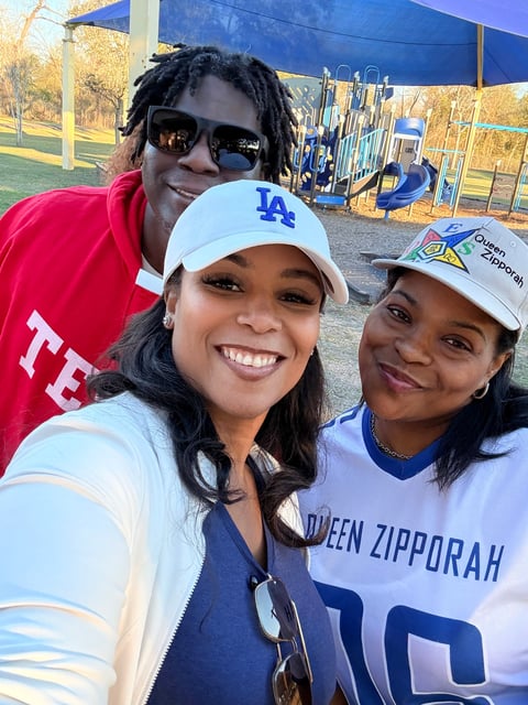 Three smiling people posing together at a park, wearing casual sports attire with blue and red colors, playground visible in background