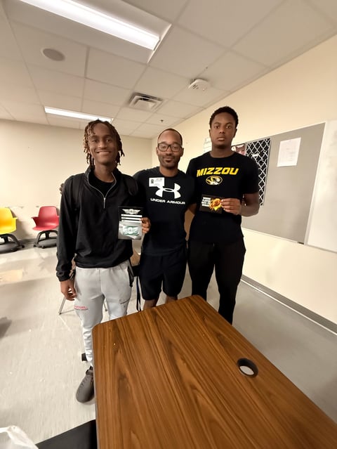 Three young men standing in an indoor room behind a wooden table, wearing black athletic clothing, in a casual indoor setting