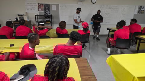 Students in red shirts sitting at yellow tables during a classroom lesson with instructors standing at the front of the room