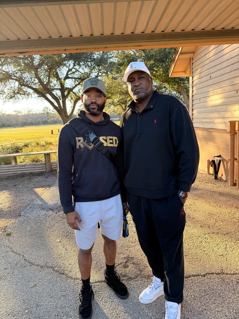 Two men standing together under a covered porch area with a rural field and trees visible behind them