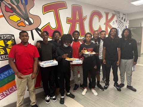 Group of people standing in front of a colorful Jack mural, some holding pizza boxes, in an indoor space