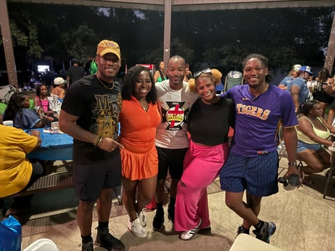Group of five smiling people posing together at an outdoor evening event with spectators visible in the background
