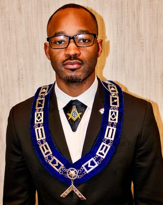 Man wearing black suit and glasses with blue Masonic regalia and emblem against beige background